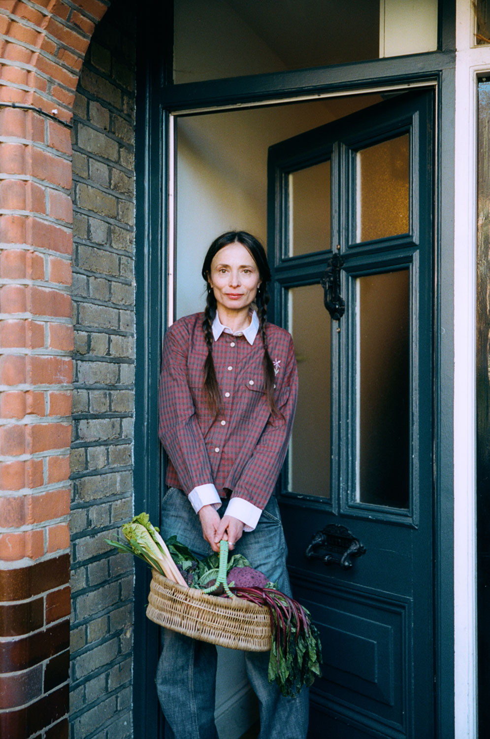 A woman stands in front of a teal blue front door, slightly ajar. She holds a wicker basket of vegetables in both hands, with her hair in plaited pigtails. She wears Saywood's Jules Utility Shirt in red micro check with contrast lilac collar and cuffs, worn with mid wash jeans.