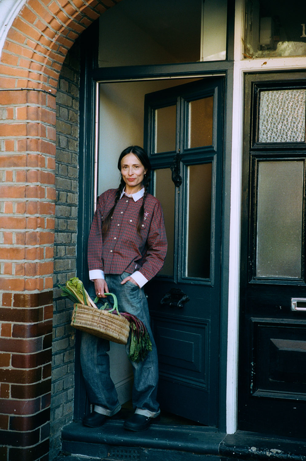 A woman stands in front of a teal blue front door, slightly ajar. She holds a wicker basket of vegetables, with her hair in plaited pigtails. She wears Saywood's Jules Utility Shirt in red micro check with contrast lilac collar and cuffs, worn with mid wash jeans, with one hand in her jean pocket.